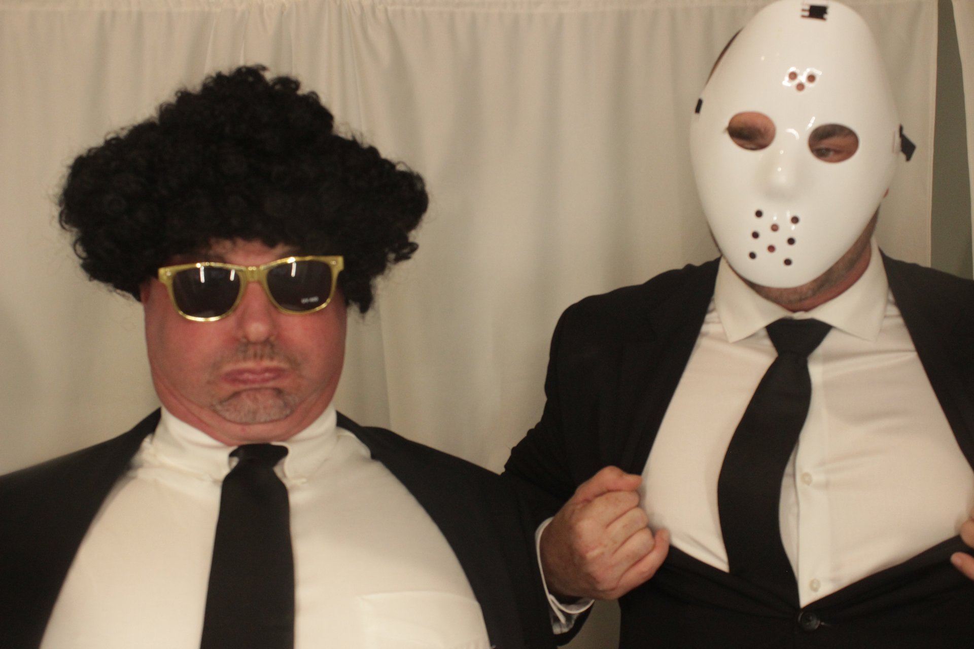 Two guests in suits posing with sunglasses, a wig, and a costume mask