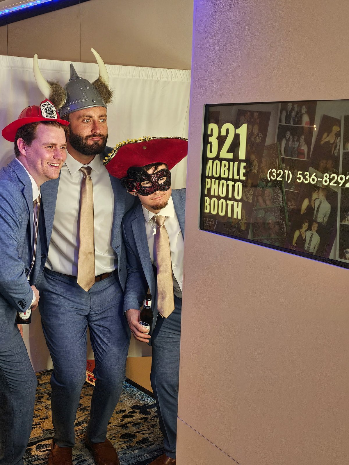 Three wedding guests posing in suits with playful hats and a mask beside the photo booth