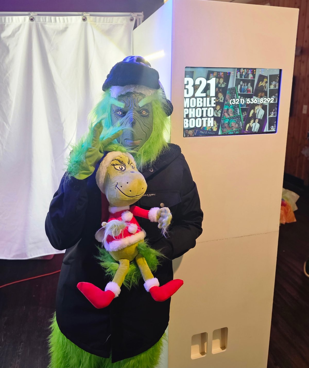 A guest in a Grinch costume holding a Grinch plush beside the photo booth